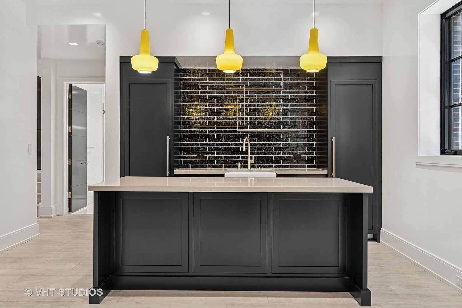 This interior shot showcases a modern kitchen with a striking black and gold backsplash, complemented by sleek black cabinetry and a light-colored countertop island. Three yellow pendant lights hang above the island, adding a pop of color and illuminating the workspace. The kitchen's design is both stylish and functional, creating an inviting space.