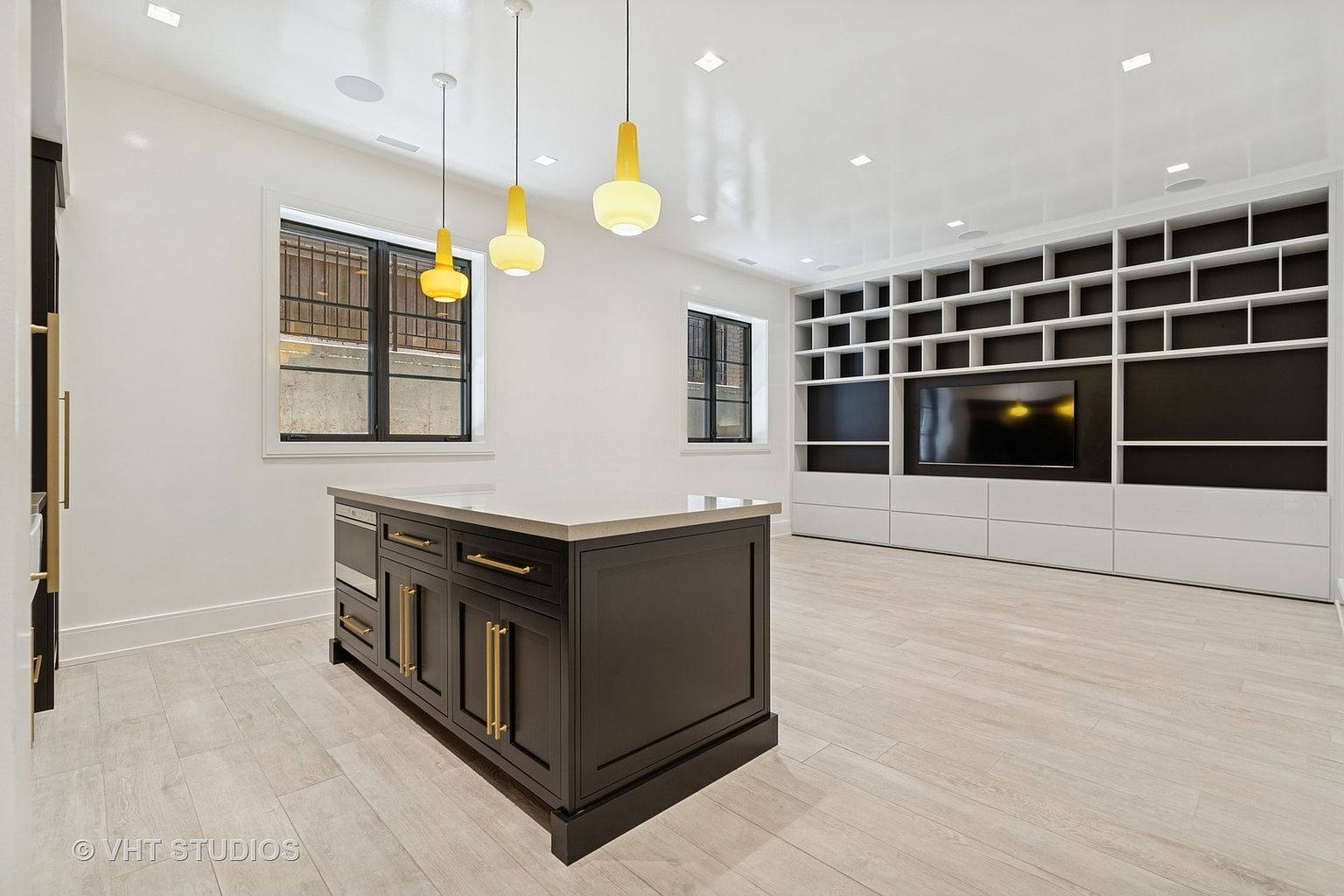This interior shot showcases a modern kitchen with a dark-toned island featuring gold hardware and a light countertop. The room is brightly lit with recessed lighting and pendant lights, complemented by natural light from two windows. A large built-in shelving unit with a television adds a unique touch to the space, creating a blend of kitchen and living area.