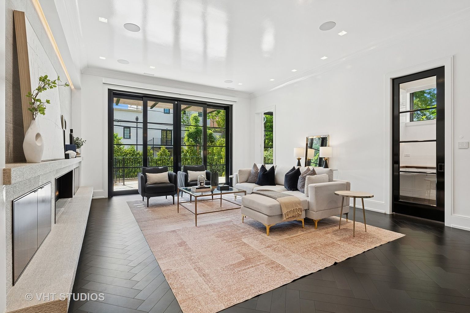 This is a bright and modern living room featuring dark herringbone wood floors, a light-colored area rug, and a neutral-toned sofa with decorative pillows. The room has large black-framed sliding glass doors leading to the outside, and a contemporary fireplace with a light-colored surround. The space is well-lit and appears spacious, creating an inviting atmosphere.
