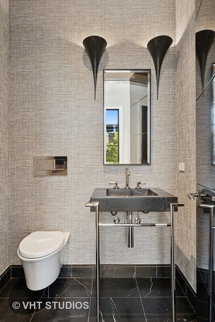 This is a stylish guest bathroom featuring a wall-mounted toilet and a unique sink with a dark stone basin and chrome stand. The walls are covered in a textured wallpaper, and the floor is black marble with white veining. Modern sconces flank a rectangular mirror, adding to the room's sophisticated aesthetic.