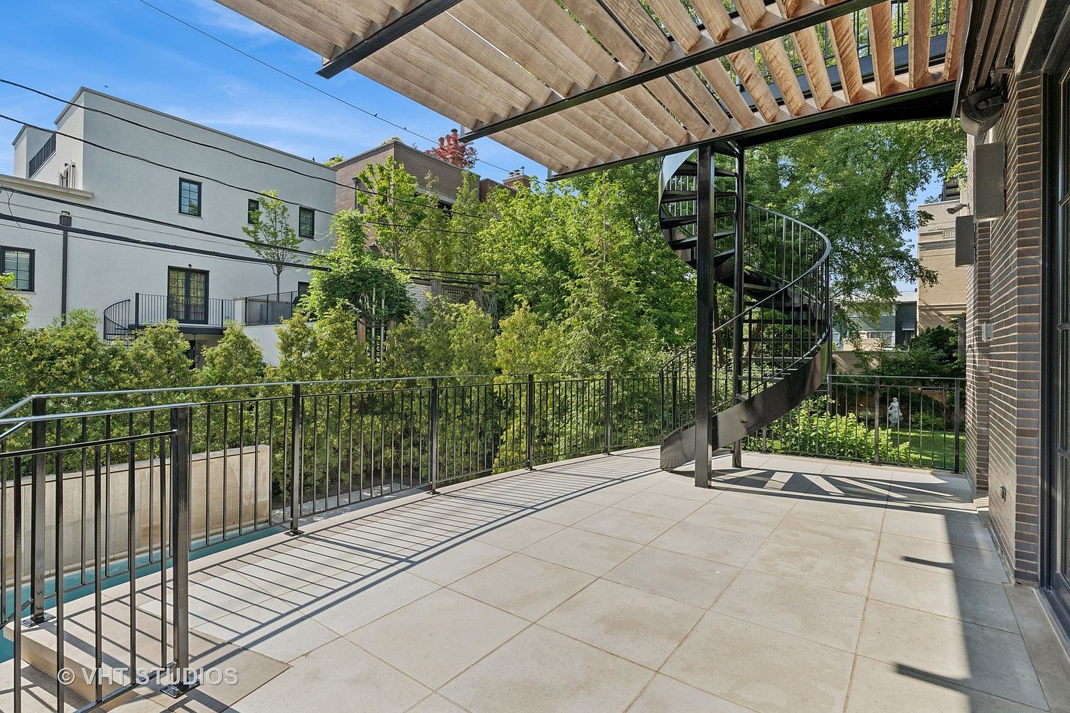 This image showcases a spacious outdoor patio or deck area, featuring a modern design with large square tiles and a black metal railing. A striking spiral staircase adds architectural interest, leading to another level. The space is partially covered by a wooden pergola, providing shade and a stylish touch, while lush greenery in the background offers privacy and a serene atmosphere.