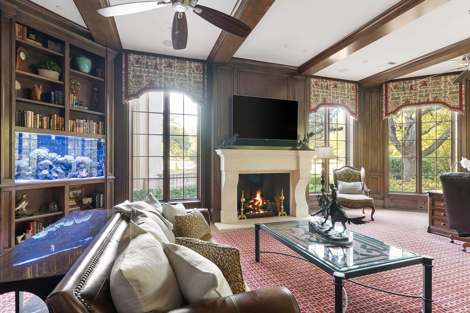 This is a luxurious living room featuring dark wood paneling, a stone fireplace with a mounted television, and custom window treatments. A large aquarium is built into the shelving unit, adding a unique focal point. The room is furnished with a leather sofa, a glass-topped coffee table with a sculpture, and a patterned rug, creating a warm and inviting atmosphere.