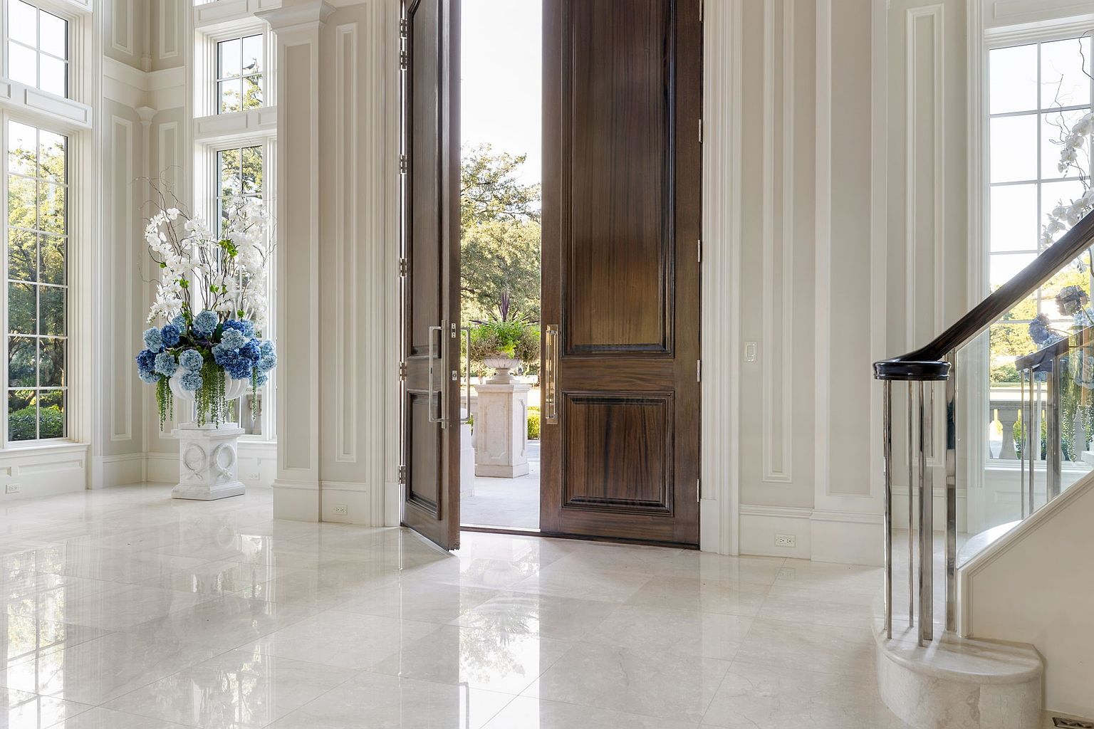 This grand foyer features a polished marble floor that reflects natural light streaming in from large windows. A striking floral arrangement adds a touch of elegance, while the open double doors invite exploration of the property. The staircase with its chrome and wood railing hints at the home's luxurious design.