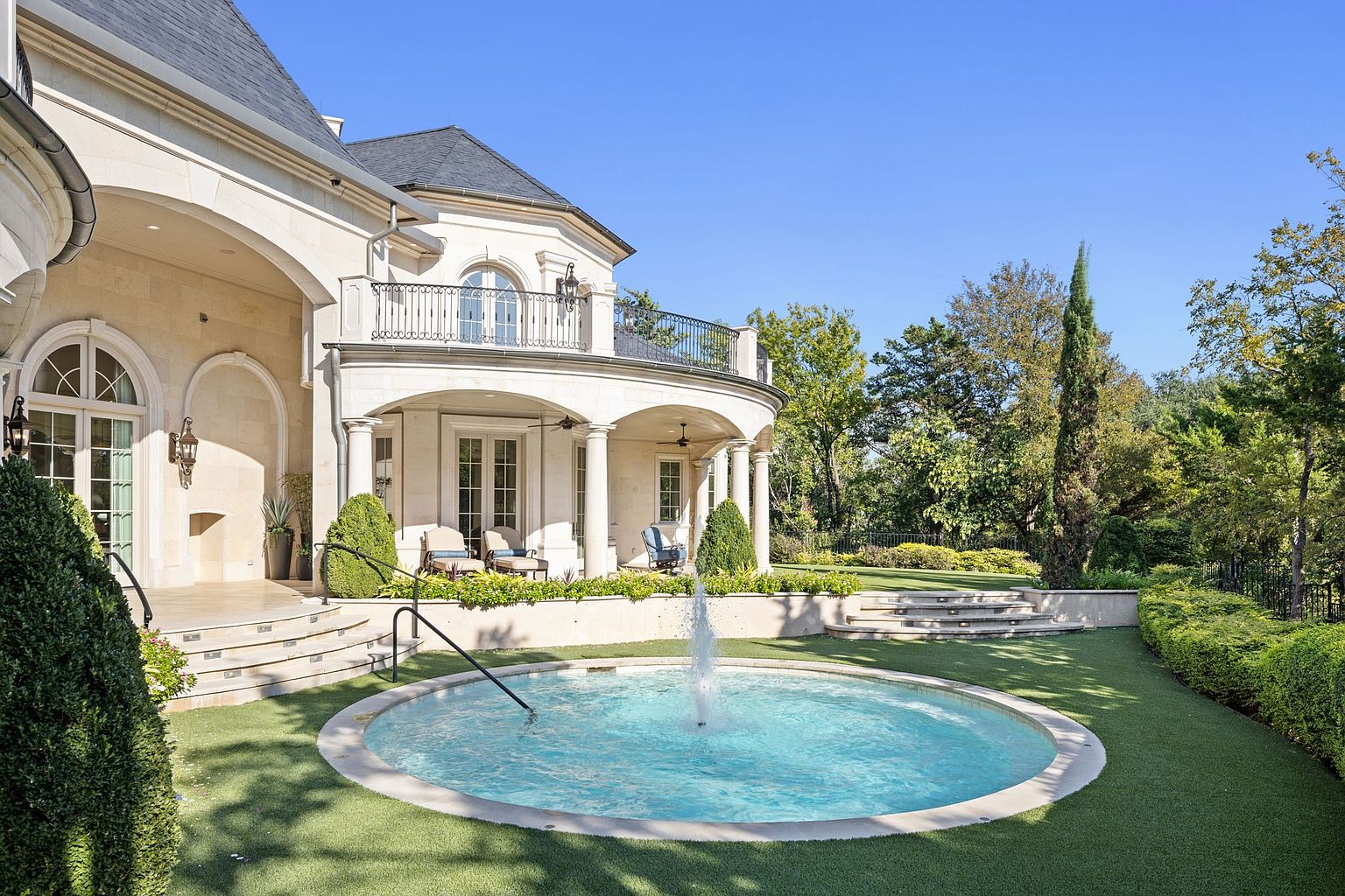 This image showcases the rear exterior of a luxurious home, featuring a stone facade, a balcony with wrought iron railings, and a manicured lawn. A circular fountain adds a focal point to the garden, while the architecture exudes elegance and sophistication. The property is surrounded by lush greenery, enhancing its privacy and appeal.