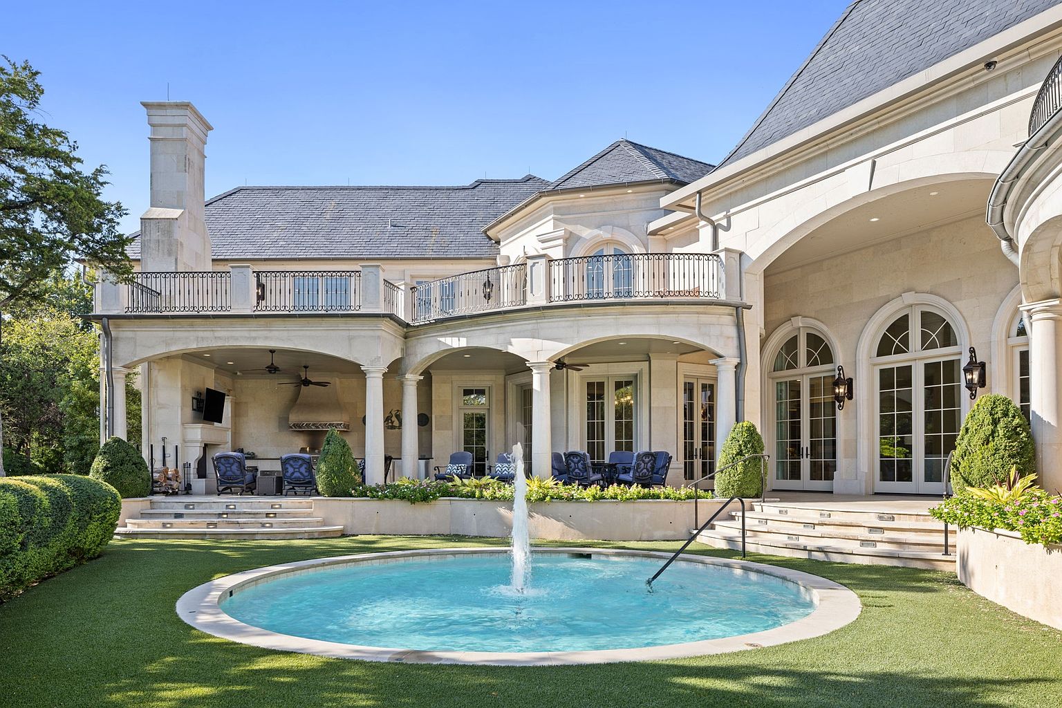 This is a rear view of a luxurious, multi-story home featuring a light-colored stone facade, a dark slate roof, and multiple balconies with wrought iron railings. A circular fountain with a central water feature is prominently displayed in the manicured lawn, adding to the property's elegance. The architecture includes arched doorways and windows, creating a sophisticated and inviting outdoor space.