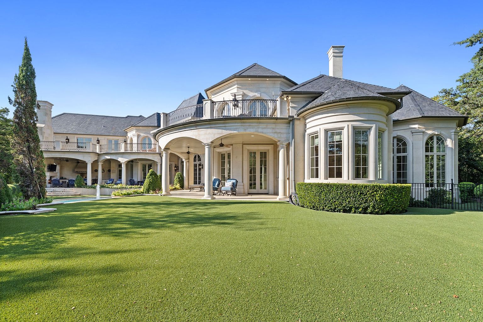 This is a rear view of a large, luxurious home featuring a manicured lawn and elegant architectural details. The house has multiple balconies, arched windows, and a covered patio area with seating. The overall impression is one of grandeur and sophisticated outdoor living.