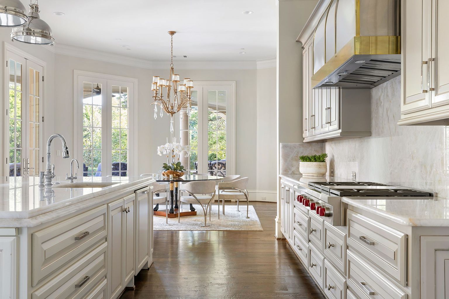 This is a bright and elegant kitchen featuring custom white cabinetry, marble countertops, and stainless steel appliances. A chandelier hangs above a glass-topped dining table with modern chairs, creating a sophisticated breakfast nook. The dark hardwood floors add warmth and contrast to the light color palette, enhancing the luxurious feel of the space.