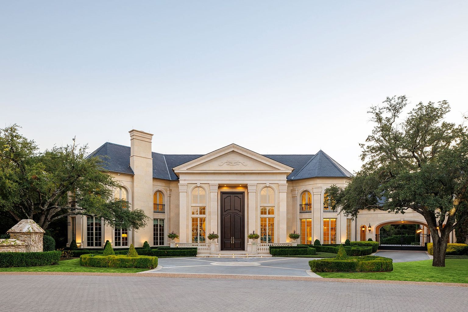 This is a grand front view of a luxurious estate, showcasing its symmetrical design, elegant stone facade, and dark roof. The manicured landscaping, including trimmed hedges and mature trees, enhances the property's curb appeal. A wide driveway leads to the impressive entrance, creating a welcoming and stately impression.