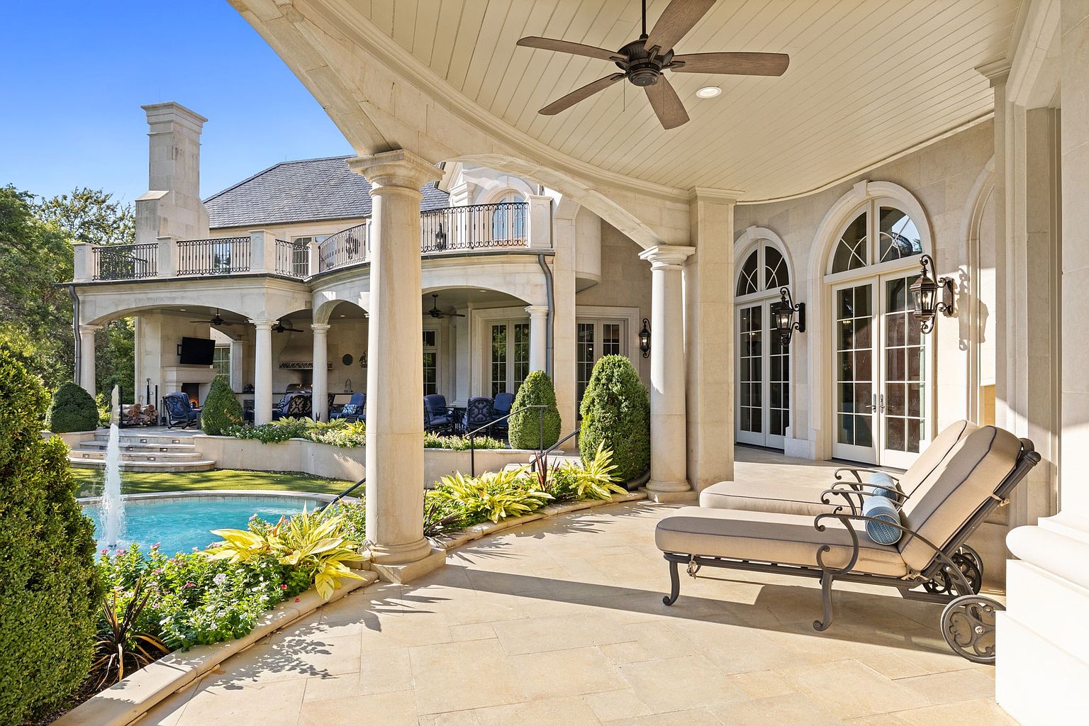 This image showcases a luxurious patio area with elegant stone columns and arches, providing a covered outdoor living space. The patio features comfortable lounge chairs, manicured landscaping, and a view of a pool with a fountain. The architectural style is sophisticated and upscale, suggesting a high-end property.