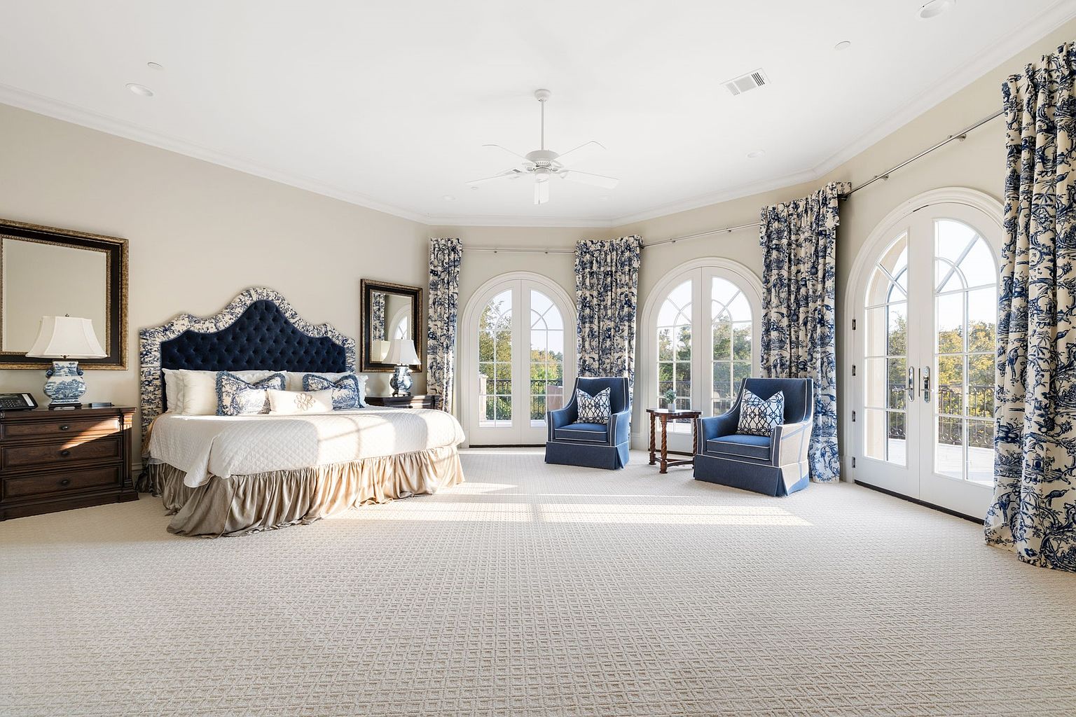 This is a spacious primary bedroom featuring a large bed with a blue upholstered headboard and matching throw pillows. The room has multiple arched windows with patterned curtains, allowing ample natural light to illuminate the patterned carpet. Two armchairs are placed near the windows, creating a cozy seating area.
