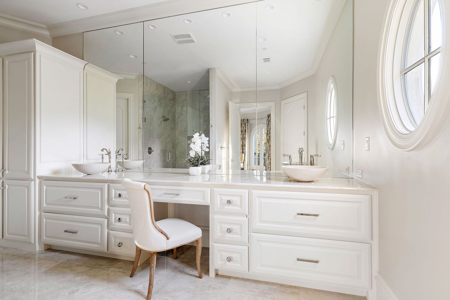 This is a bright and luxurious primary bathroom featuring a double vanity with marble countertops and vessel sinks. The large mirror enhances the sense of space, reflecting the natural light from the oval window. The white cabinetry and neutral color palette create a clean and elegant aesthetic.