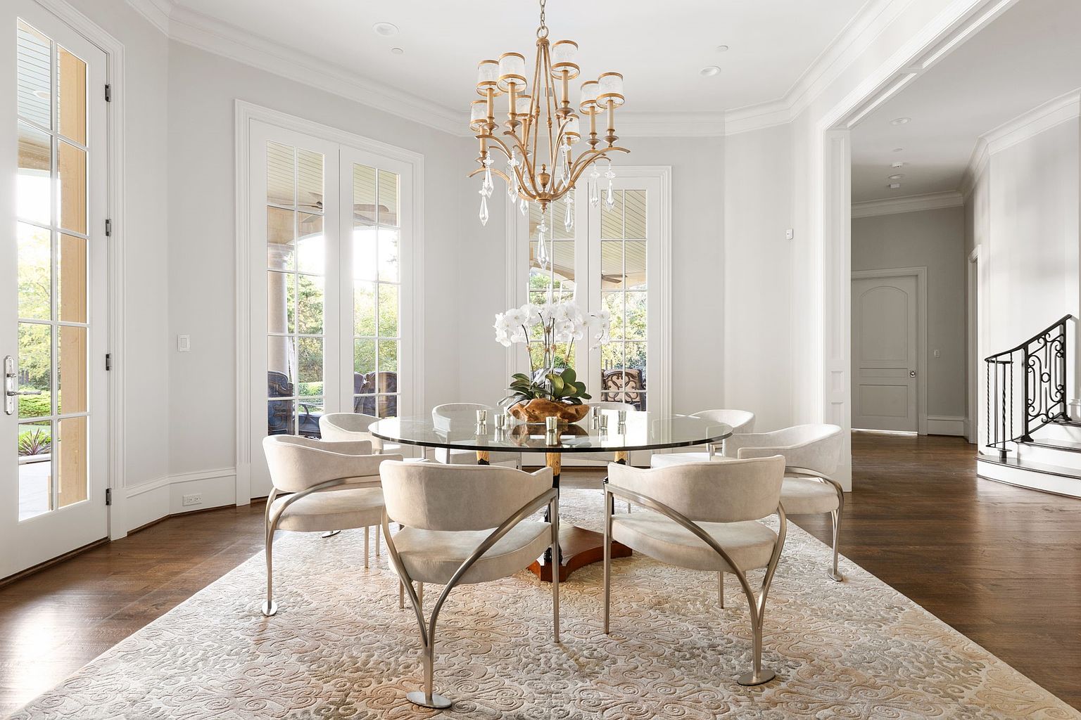 This is an interior shot of a dining room featuring a round glass table with six chairs. A gold chandelier hangs above the table, which is adorned with a floral centerpiece. The room is bright and airy, with large windows and a neutral color palette, creating an elegant and inviting atmosphere.