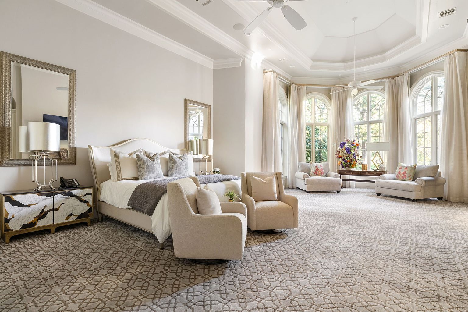 This is a luxurious primary bedroom featuring a large bed with decorative pillows and a gray throw blanket. The room is spacious with multiple seating areas, including armchairs near large arched windows draped with sheer curtains. The neutral color palette and patterned carpet create a serene and elegant atmosphere.