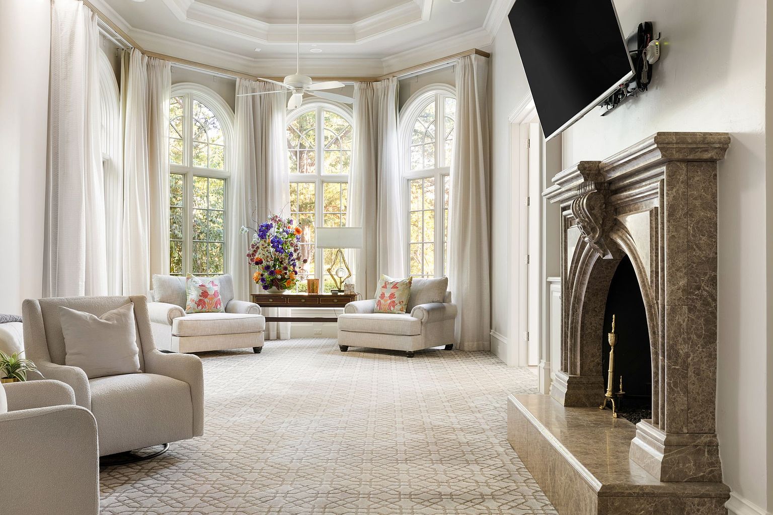 This is a bright and airy living room featuring a bay window with sheer white curtains, allowing natural light to flood the space. The room is furnished with comfortable armchairs and a marble fireplace with a mounted television above. The neutral color palette and patterned rug create a sophisticated and inviting atmosphere.