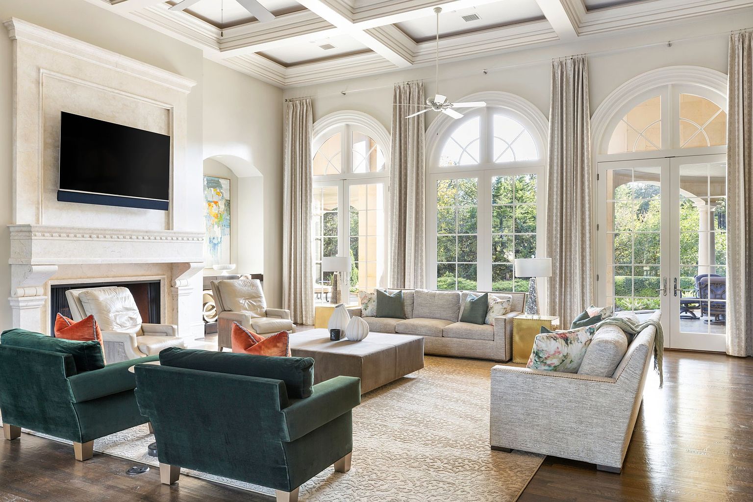 This is a spacious and luxurious living room featuring high ceilings with coffered detailing, large arched windows with elegant drapery, and a sophisticated color palette. The room is furnished with comfortable seating arrangements, including sofas and armchairs, arranged around a central coffee table and a fireplace with a mounted television. The overall impression is one of refined elegance and comfortable living.