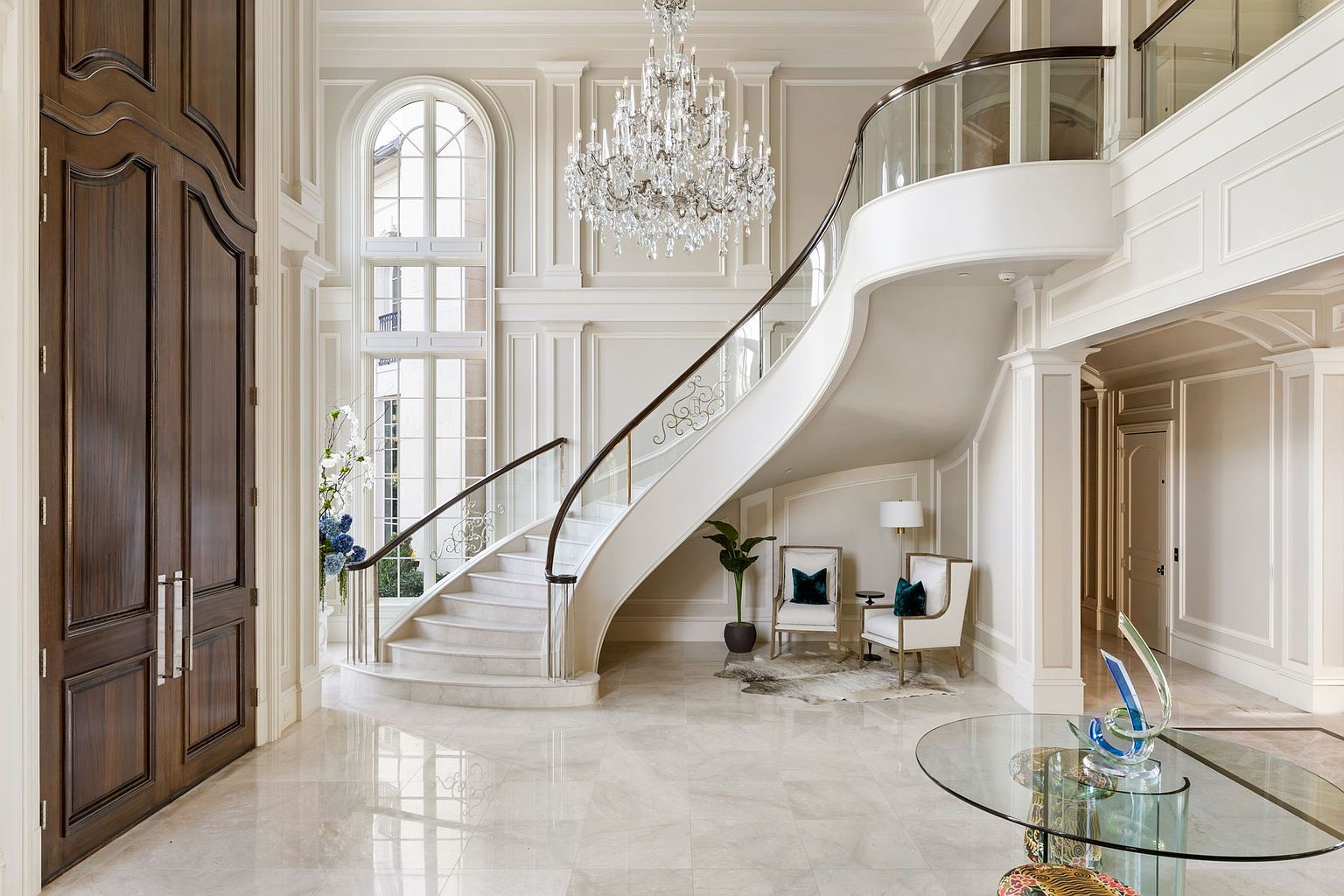 This grand foyer features a sweeping staircase with a glass and wood railing, illuminated by a large crystal chandelier. The space is decorated in neutral tones with detailed wall paneling and marble flooring, creating an elegant and luxurious atmosphere. A large arched window provides natural light, enhancing the spacious feel of the entryway.