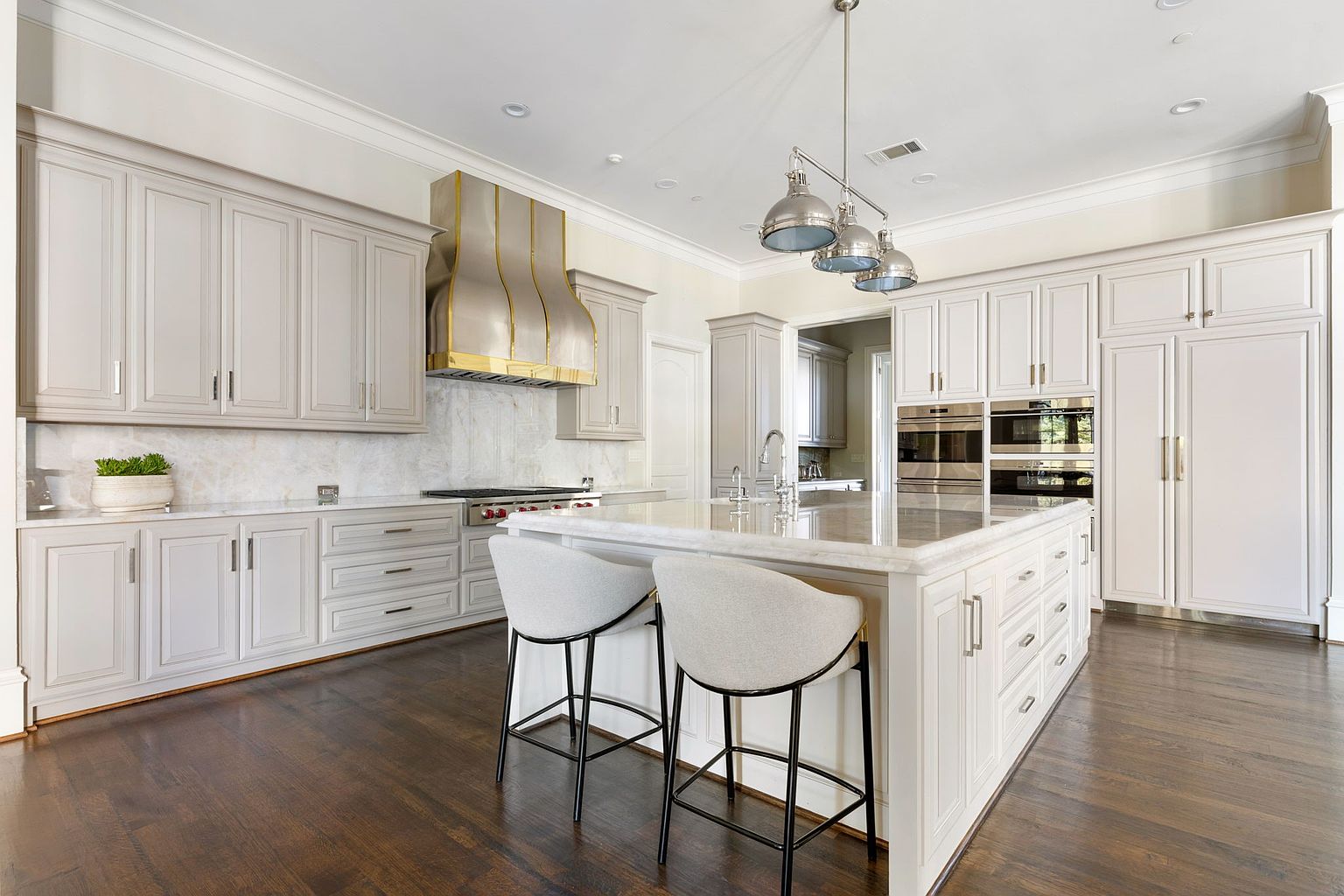 This is a bright and spacious kitchen featuring a large central island with bar seating, custom cabinetry, and high-end stainless steel appliances. The kitchen is well-lit with pendant lighting and recessed lights, and the dark hardwood floors provide a nice contrast to the light-colored cabinets. The overall impression is one of luxury and functionality.