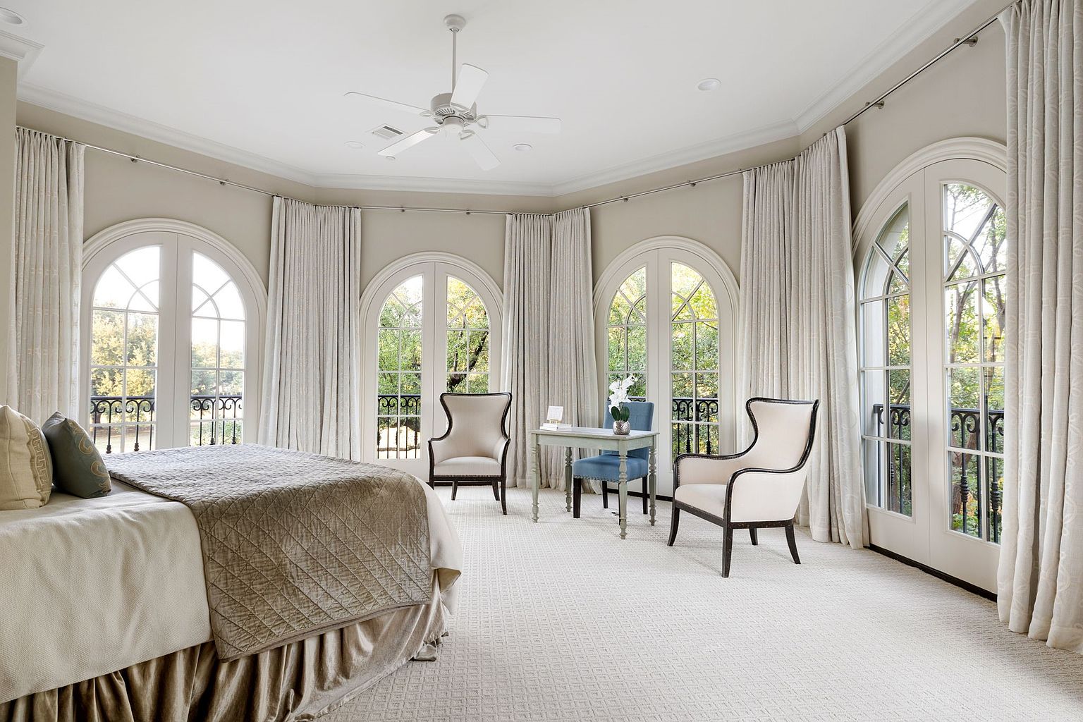 This is a spacious primary bedroom featuring large arched windows with views of the outdoors, complemented by elegant curtains. The room includes a bed with a neutral-toned comforter and a small desk area with chairs, creating a serene and luxurious atmosphere. A ceiling fan provides air circulation, and the light-colored carpet adds to the room's brightness.