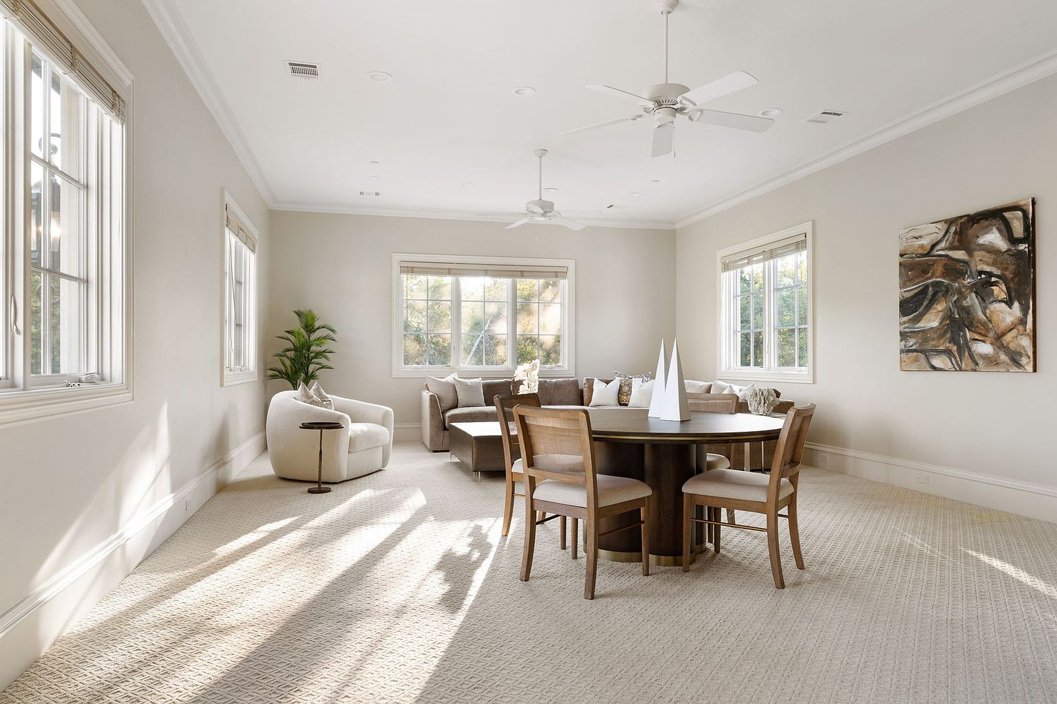 This is a bright and spacious living room featuring neutral tones and ample natural light. The room includes a comfortable seating area with a sectional sofa, a round dining table with chairs, and an accent chair with a side table. The walls are adorned with artwork, and the overall impression is one of comfort and sophistication.