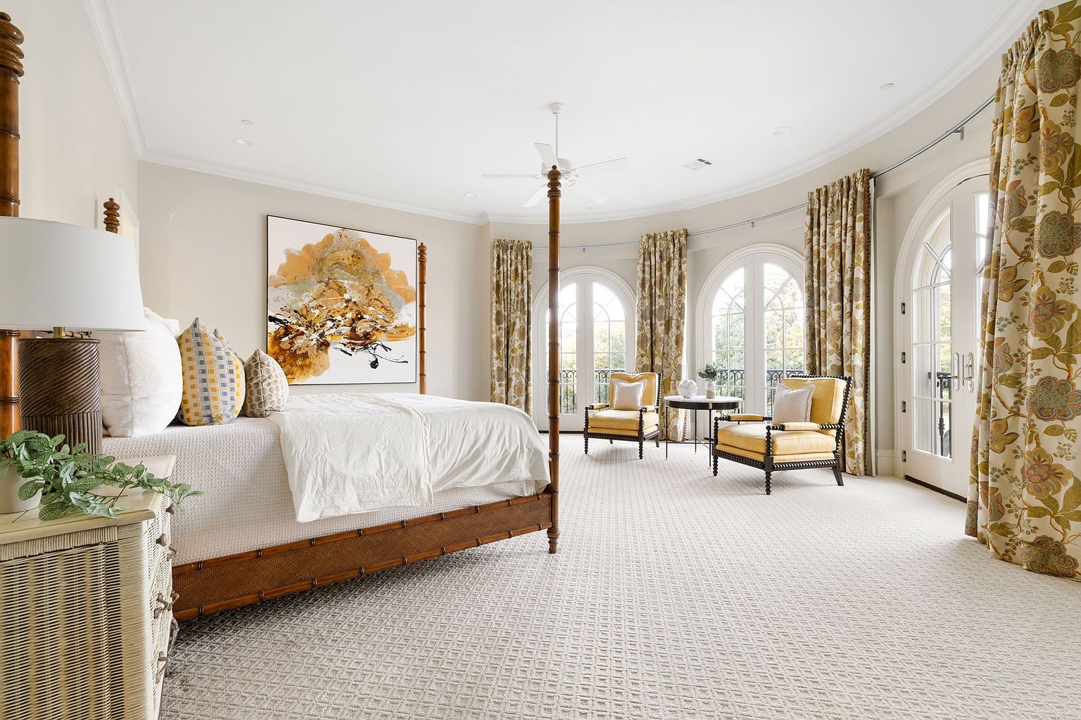 This is a spacious primary bedroom featuring a four-poster bed with luxurious bedding, complemented by an abstract art piece above the headboard. The room boasts a curved wall with large arched windows draped with patterned curtains, allowing ample natural light to illuminate the patterned carpet and seating area with two armchairs and a small table. The overall impression is one of elegance and comfort.