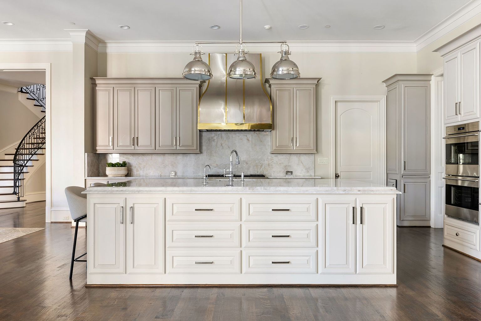 This is a well-lit kitchen featuring a large island with white cabinetry and drawers, topped with a marble countertop. The kitchen also includes gray upper cabinets, stainless steel appliances, and a custom range hood with gold accents. The hardwood floors and neutral color palette create a sophisticated and inviting atmosphere.