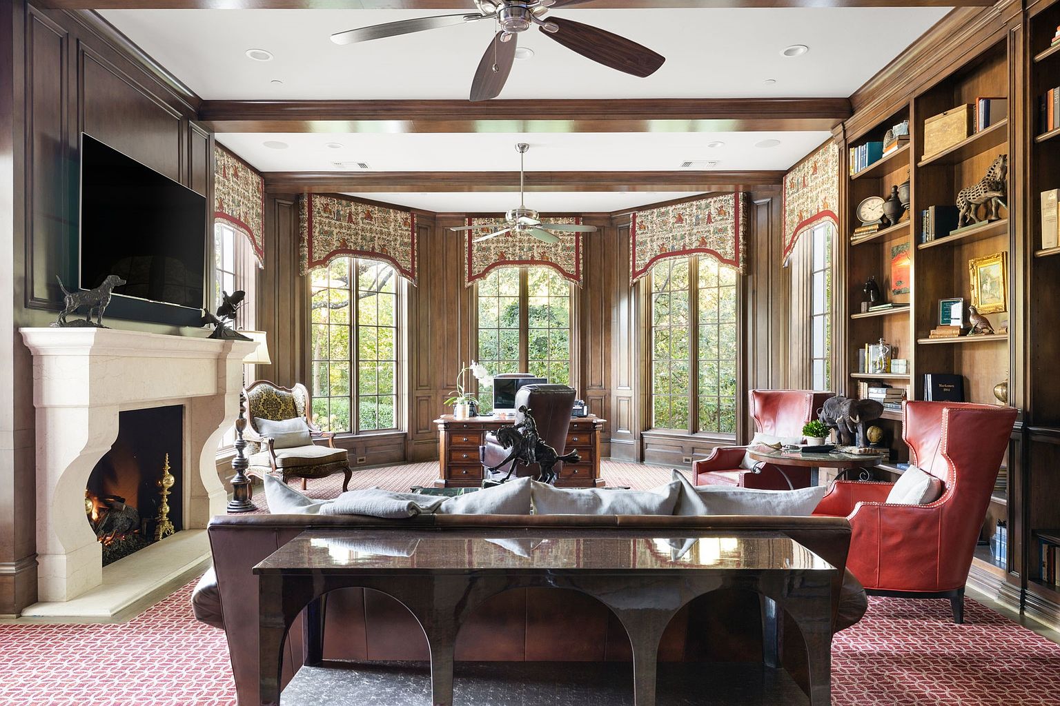 This is a luxurious home office featuring rich wood paneling, a large bay window with patterned valances, and custom built-in bookshelves. A stone fireplace with a mounted television adds a focal point, while leather seating and a patterned rug contribute to the room's sophisticated and comfortable atmosphere. The space exudes a sense of classic elegance and professional refinement.