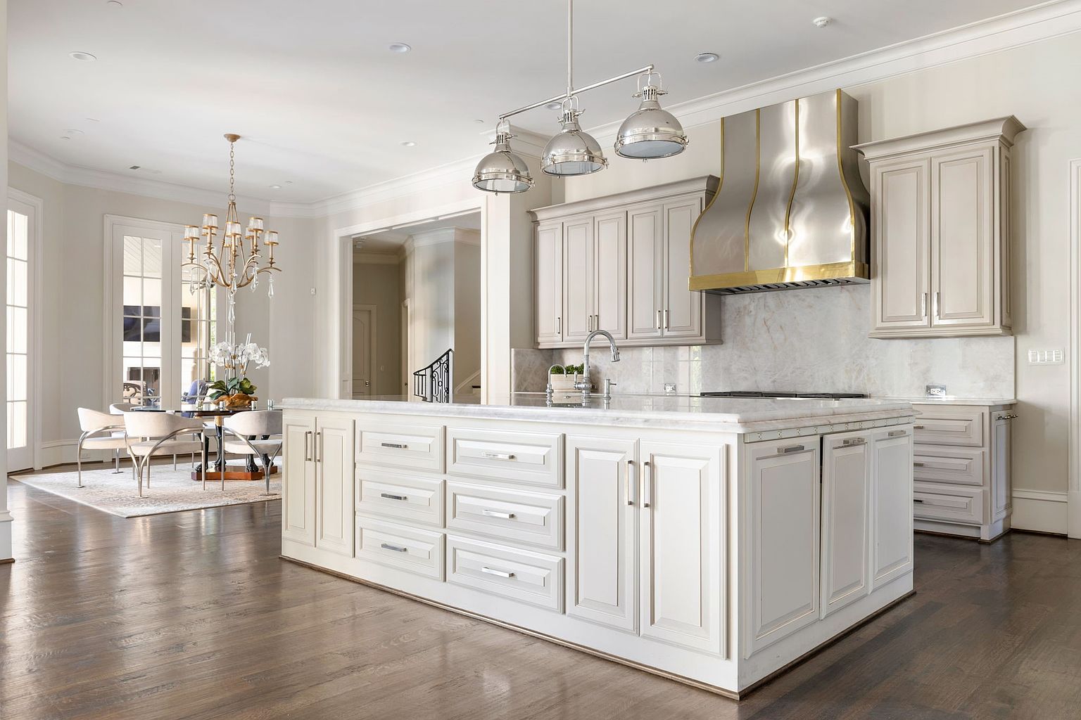 This is a bright and spacious kitchen featuring a large central island with white cabinetry and marble countertops. Stainless steel pendant lights hang above the island, complementing the stainless steel range hood. The kitchen seamlessly transitions into a dining area with a chandelier, creating an open and inviting space.