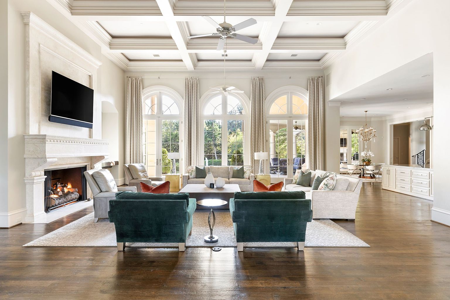 This is a spacious and luxurious living room featuring a high coffered ceiling with a ceiling fan, a fireplace with a large television mounted above, and three large arched windows providing ample natural light. The room is furnished with multiple sofas and armchairs, arranged around a central coffee table, creating a comfortable and inviting atmosphere. The dark hardwood floors contrast beautifully with the light-colored walls and furniture, adding to the room's elegance.
