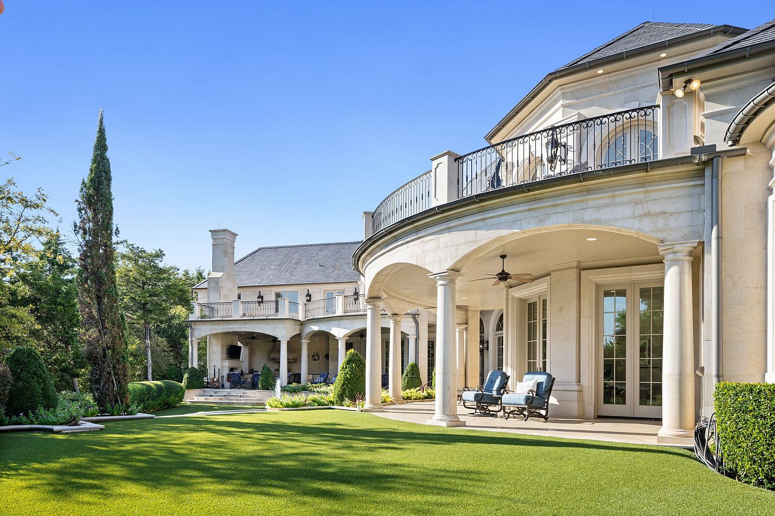This is a rear view of a luxurious estate, showcasing a sprawling green lawn and an elegant stone facade. The architecture features multiple balconies with wrought iron railings, arched colonnades, and large windows, creating a sophisticated and inviting outdoor living space. The manicured landscaping and architectural details emphasize the property's high-end appeal.