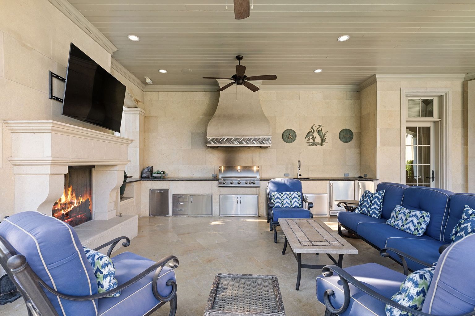 This is a luxurious outdoor patio area featuring a full outdoor kitchen with stainless steel appliances, a stone fireplace with a mounted television, and comfortable blue cushioned seating. The space is well-lit with recessed lighting and a ceiling fan, creating an inviting atmosphere for relaxation and entertainment. The flooring is tiled and the walls are stone.