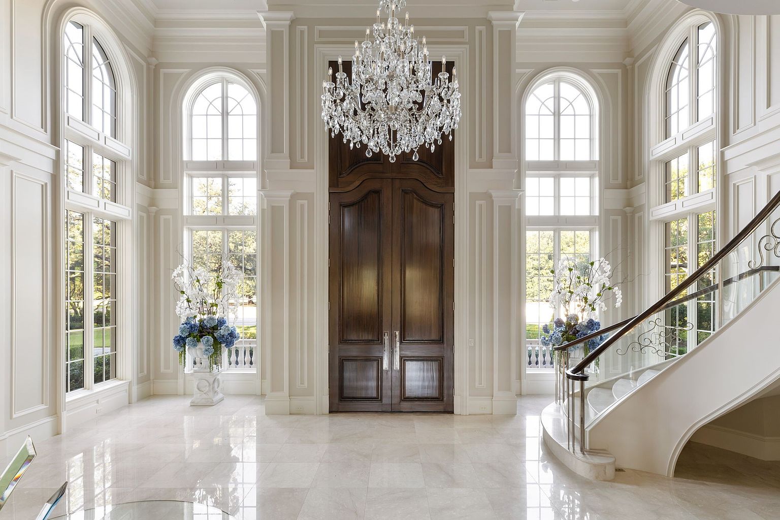 This grand foyer features a soaring ceiling with an ornate crystal chandelier, a sweeping staircase with wrought iron detailing, and large arched windows that flood the space with natural light. The elegant marble flooring and custom millwork contribute to the luxurious and sophisticated ambiance, creating a memorable first impression. A large wooden double door is centered in the back.