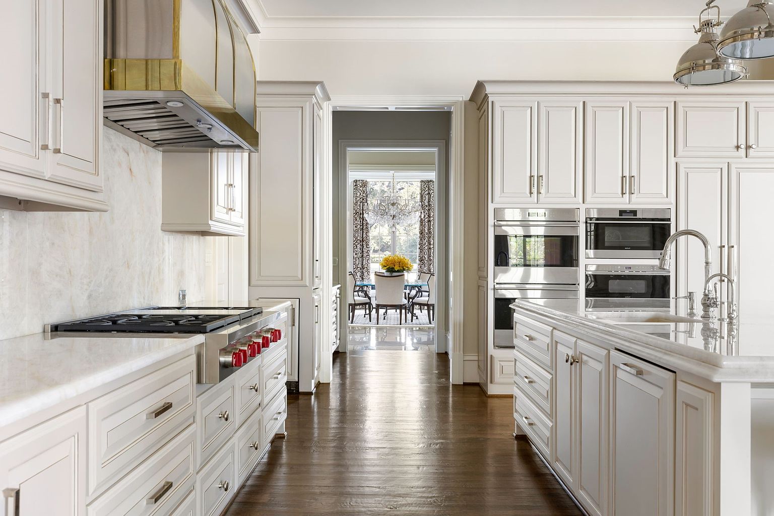 This is a bright and elegant kitchen featuring white cabinetry, stainless steel appliances, and a marble countertop. The kitchen is well-lit and spacious, with a view into the dining room. The dark hardwood floors add a touch of warmth and sophistication to the space.