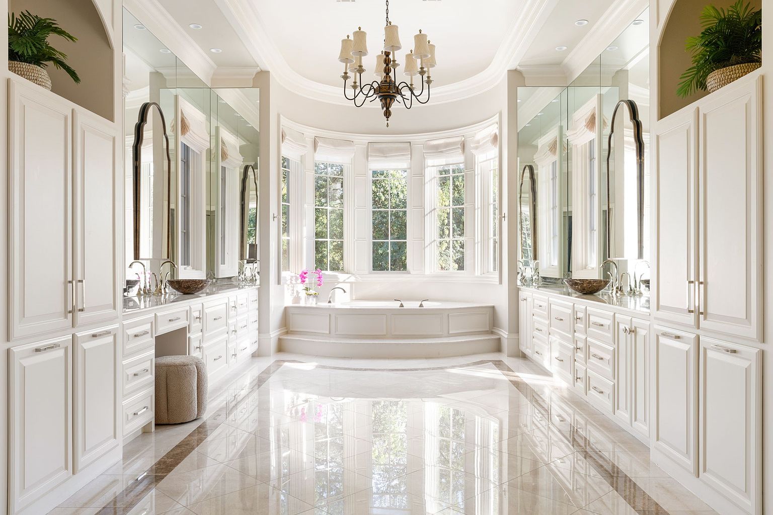 This is a luxurious primary bathroom featuring dual vanities with white cabinetry, large mirrors, and elegant fixtures. A central soaking tub is positioned beneath a bay window, offering natural light and a focal point. The marble flooring and chandelier add to the room's opulent and spacious feel.