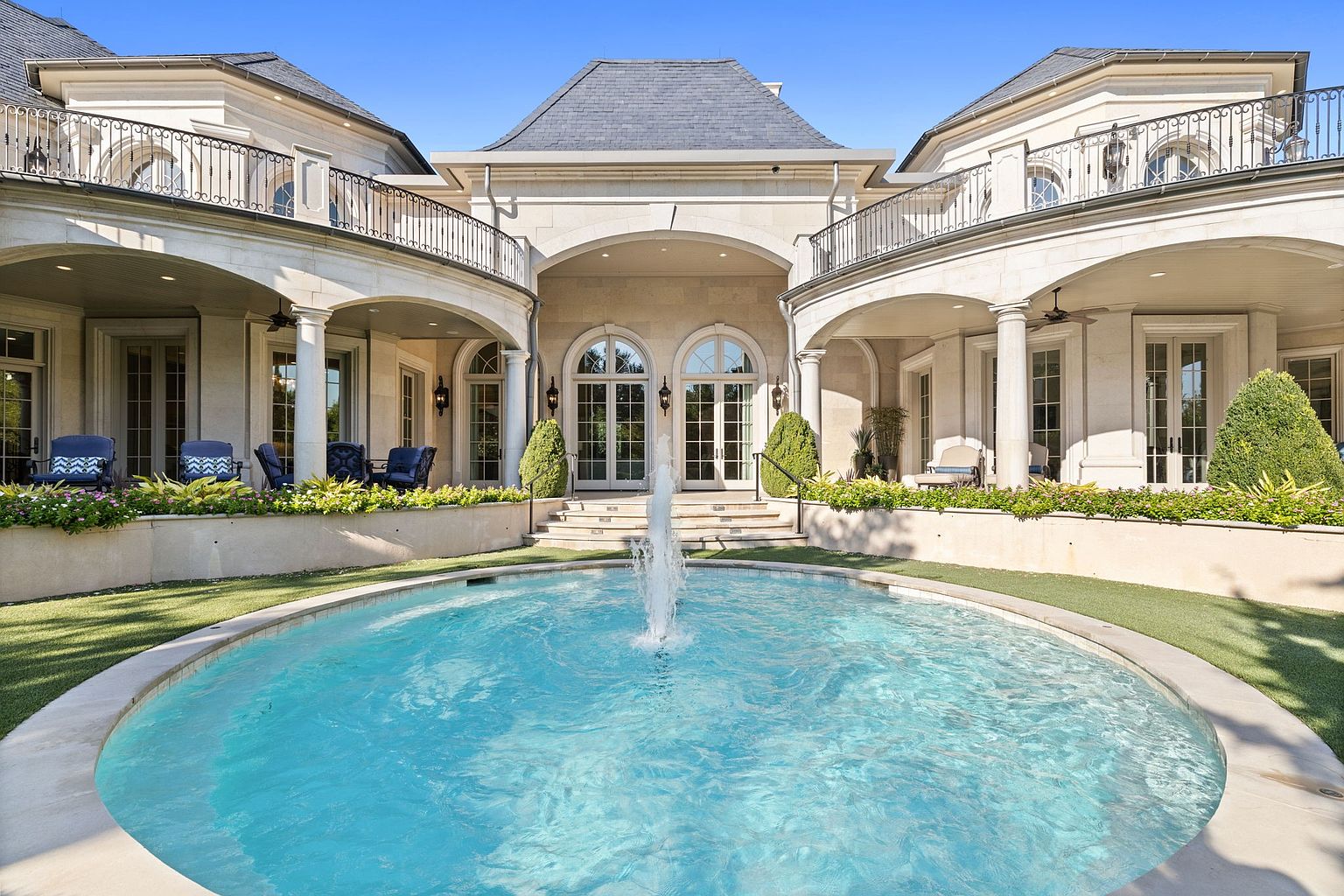 This image showcases the rear exterior of a luxurious home, focusing on a circular pool with a central fountain. The architecture features elegant stone construction, arched doorways, and wrought-iron balconies. Lush landscaping surrounds the pool, creating a serene and upscale ambiance.