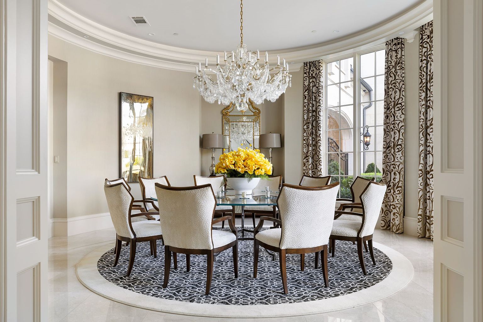 This is an elegant dining room featuring a round glass table surrounded by upholstered chairs with dark wood frames, all set upon a patterned circular rug. A large crystal chandelier hangs above the table, and the room is decorated with mirrors and lamps, creating a luxurious and inviting atmosphere. The room is bathed in natural light from the large windows, which are framed by patterned curtains.