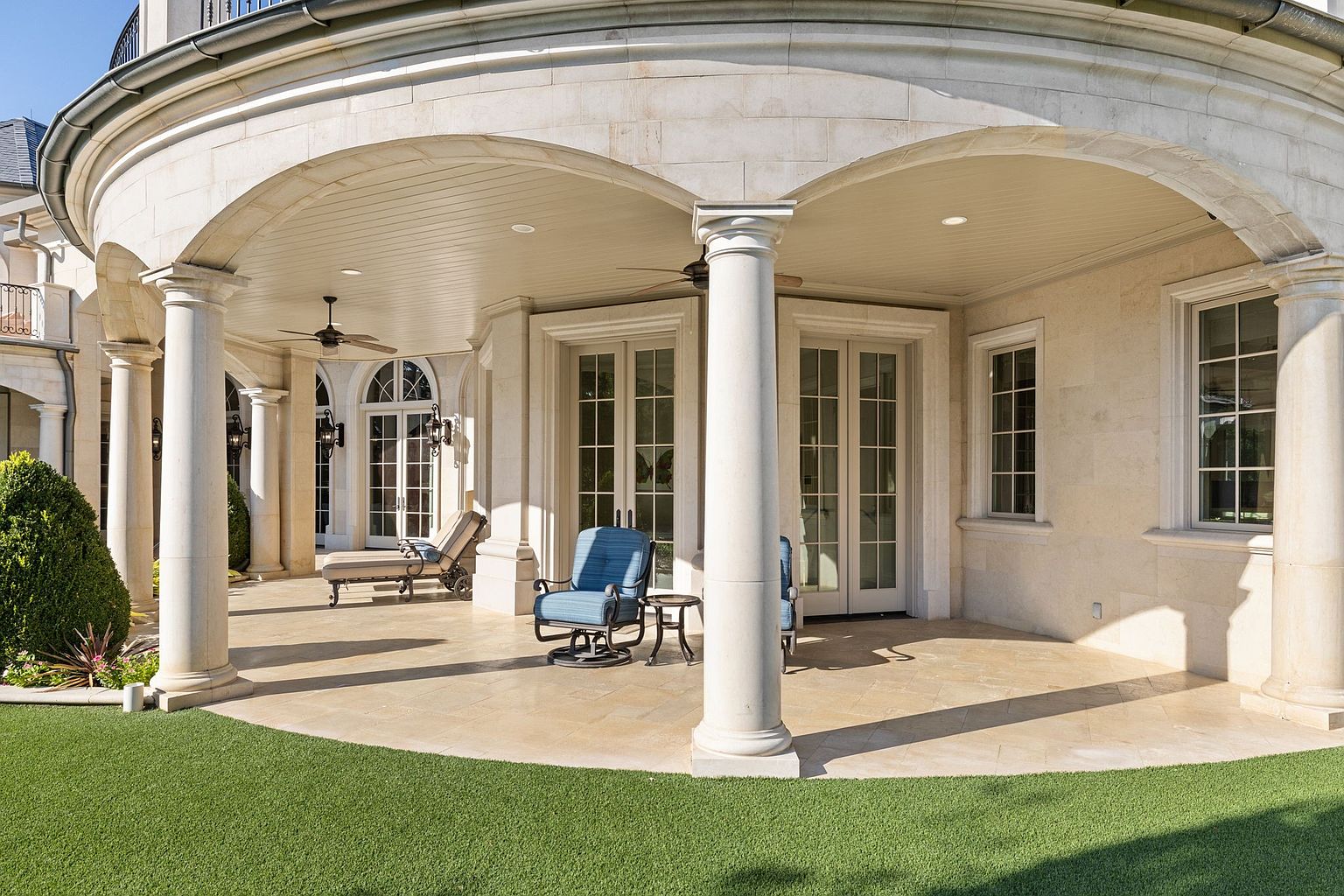 This image showcases a luxurious outdoor patio area, featuring elegant stone columns and arches that provide a covered space. The patio is furnished with comfortable seating, including blue chairs and a chaise lounge, inviting relaxation. The manicured lawn extends from the patio, enhancing the overall appeal of this outdoor living space.
