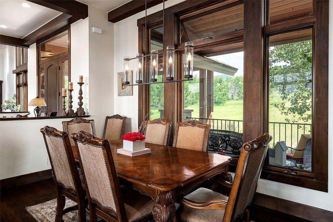 This is an interior shot of a dining room featuring a large wooden dining table with upholstered chairs. A modern chandelier hangs above the table, and large windows offer a view of the outdoor landscape. The room exudes a sense of rustic elegance and comfort, making it ideal for family gatherings or formal dinners.