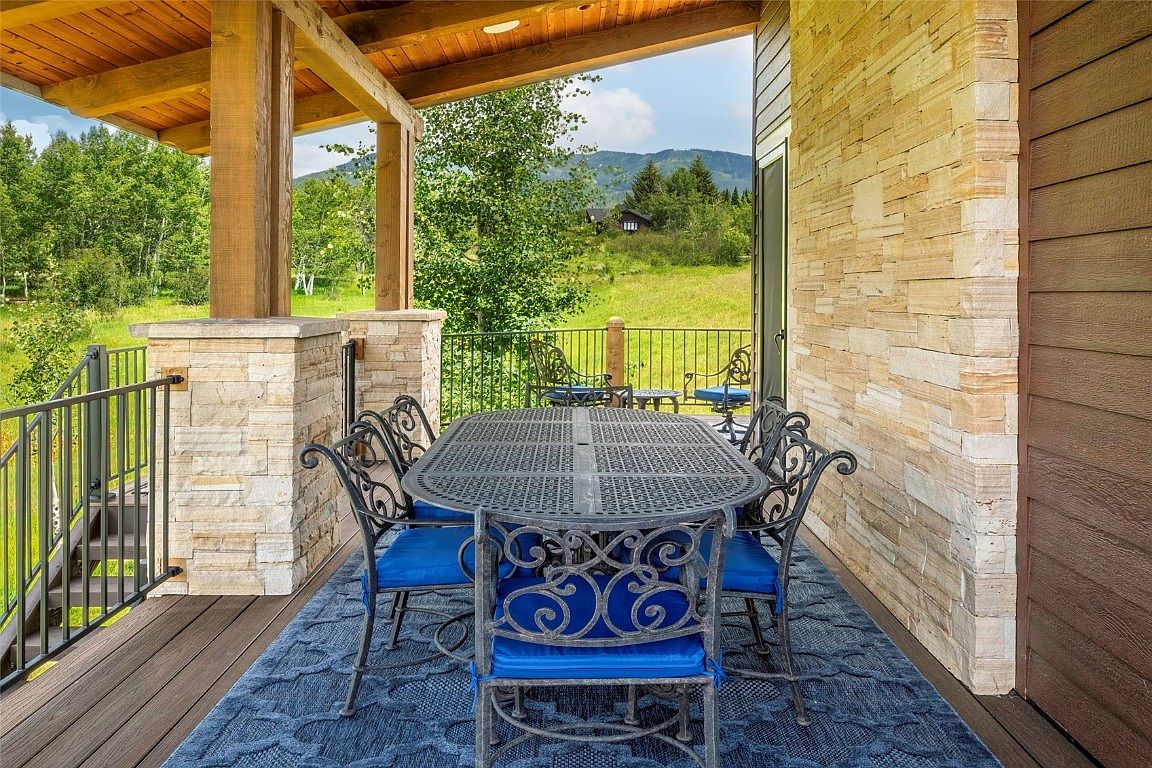 This image showcases an inviting outdoor patio area, furnished with a wrought iron dining set featuring blue cushions, set atop a blue rug. The patio is partially covered by a wooden structure supported by stone pillars, offering a view of a lush green lawn and distant mountains. The overall impression is one of comfortable outdoor living and relaxation.