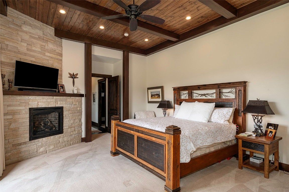 This is a primary bedroom featuring a stone fireplace with a mounted television, a large wooden bed with a patterned comforter, and a wooden ceiling with exposed beams and recessed lighting. The room has a rustic yet luxurious feel, with warm tones and natural materials creating a cozy and inviting atmosphere. The perspective is from the doorway, showcasing the entire room.