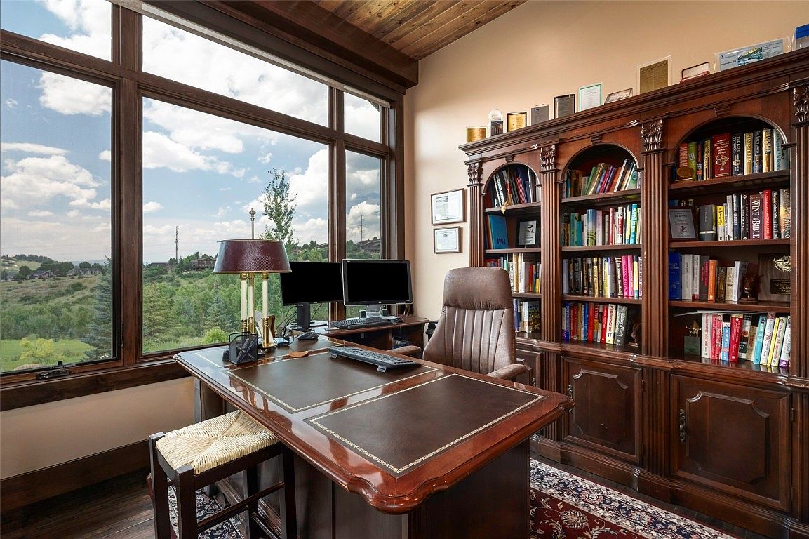This is an interior shot of a luxurious home office. The room features a large wooden desk with a leather inlay, a comfortable leather chair, and a substantial wooden bookcase filled with books. A large window offers a view of the landscape outside, creating a bright and inviting workspace.