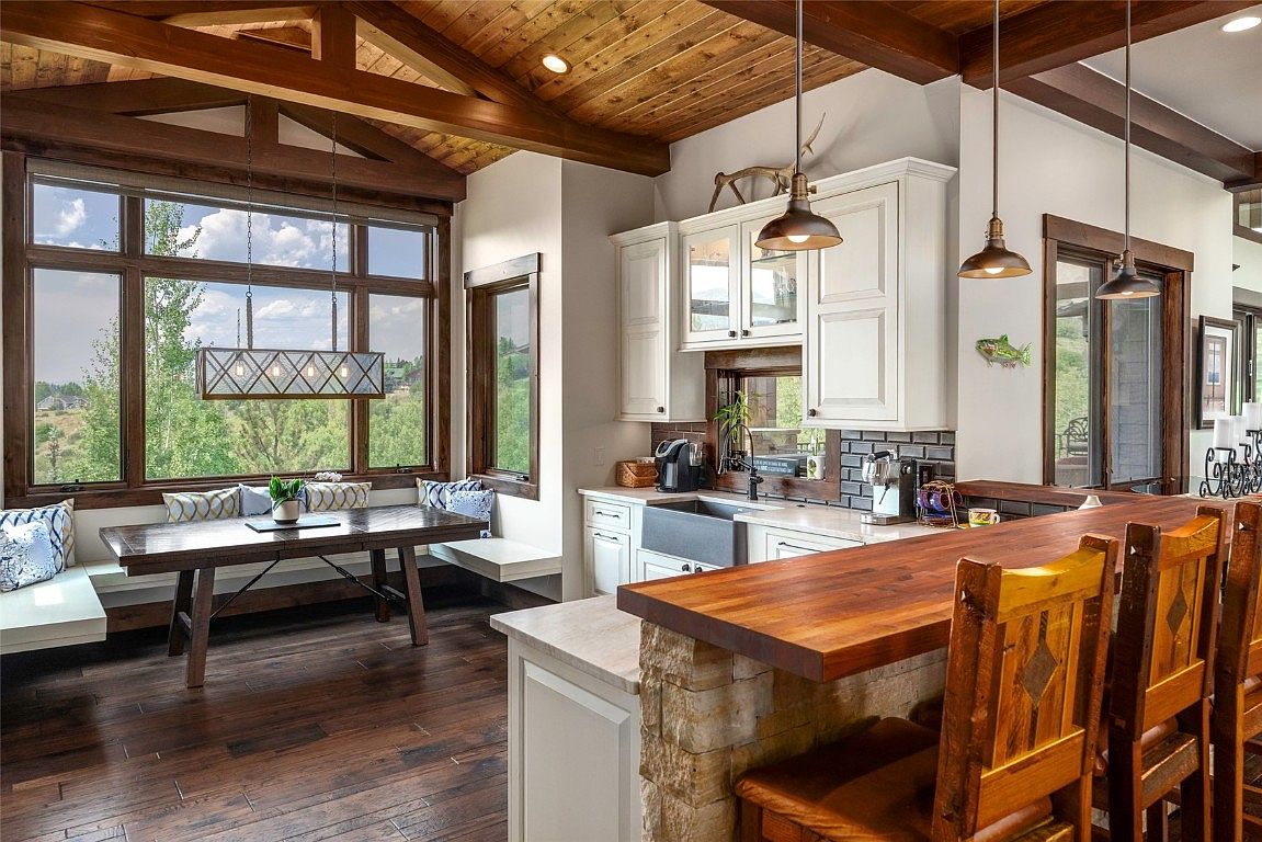This interior shot showcases a well-lit kitchen and dining area with a rustic-modern aesthetic. The kitchen features white cabinetry, a farmhouse sink, and a wooden countertop bar with stone accents, complemented by wooden bar stools. The dining area boasts a large window offering a scenic view, a wooden table, and built-in bench seating, all under a wooden beam ceiling, creating a warm and inviting atmosphere.