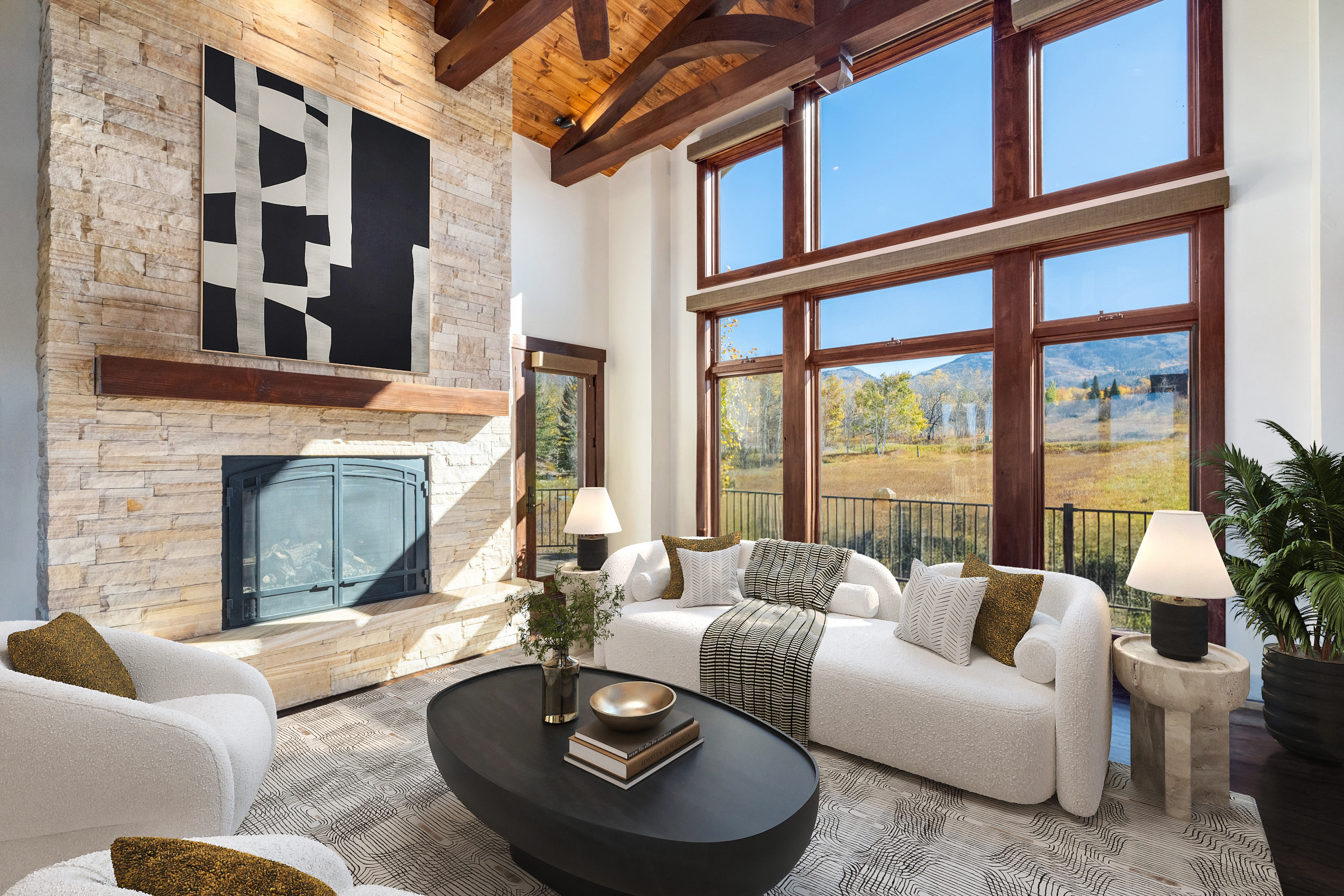 The living room showcases a stone fireplace with a black and white abstract painting above it. Large windows frame a picturesque outdoor view, complemented by the room's high, wood-beamed ceiling. The space is furnished with a chic, curved white sofa and a modern coffee table, creating an inviting and luxurious atmosphere.