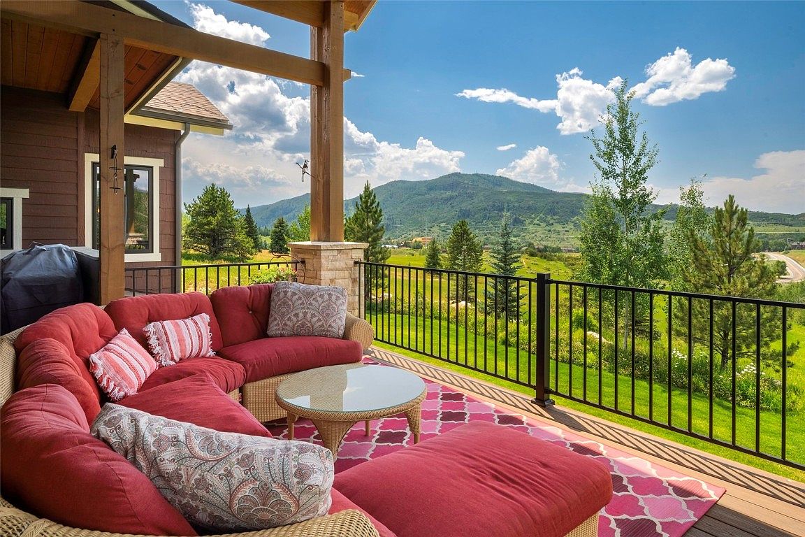 This image showcases a beautifully furnished outdoor patio or deck area, featuring a comfortable red sectional sofa with decorative pillows, a round glass-topped coffee table, and a matching ottoman on a patterned rug. The space is framed by a black metal railing, offering stunning views of a lush green landscape with rolling hills and a clear blue sky with scattered clouds. The overall impression is one of luxury and relaxation, perfect for enjoying the outdoors.