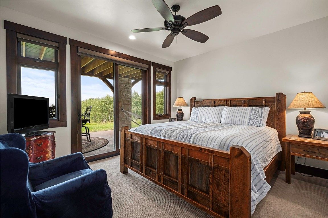 This is a primary bedroom featuring a large wooden bed with striped bedding, flanked by matching nightstands and lamps. A sliding glass door leads to an outdoor patio, offering natural light and a view of the landscape. The room also includes a comfortable blue armchair and a television, creating a cozy and inviting atmosphere.
