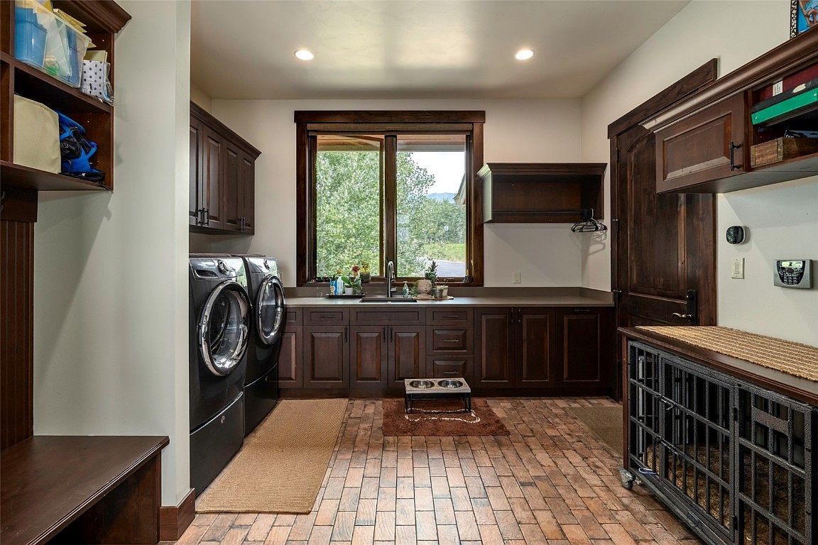 This is a well-appointed laundry room featuring dark wood cabinetry, a countertop workspace, and a window providing natural light. Modern black front-loading washer and dryer units are present, along with a dog kennel and a dog bowl station, suggesting a pet-friendly design. The brick flooring adds a touch of rustic charm to the space.