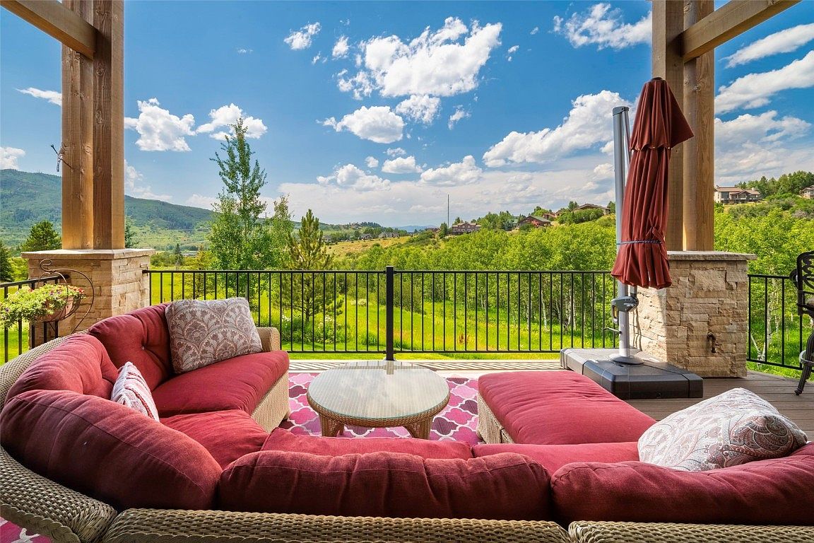 This image showcases a luxurious outdoor patio or deck area, featuring comfortable wicker seating with plush red cushions and decorative pillows. A round glass-topped table sits on a patterned rug, and a partially folded umbrella provides shade. The space overlooks a scenic landscape of rolling hills and lush greenery, creating an inviting and relaxing atmosphere.