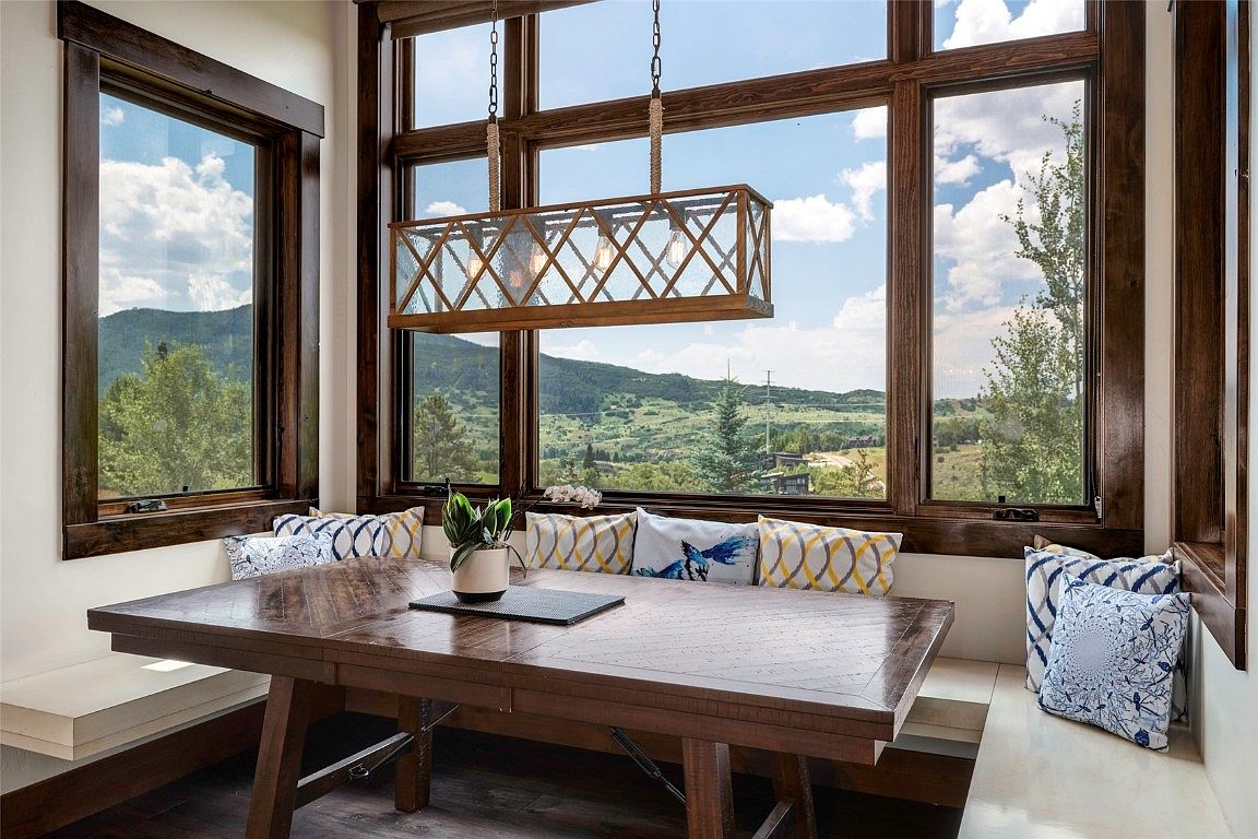 This interior shot showcases a dining area with a large wooden table set against a backdrop of expansive windows offering scenic mountain views. The room is illuminated by a unique, rectangular chandelier, and built-in seating with decorative pillows adds comfort and style. The dark wood trim around the windows complements the table, creating a warm and inviting atmosphere.
