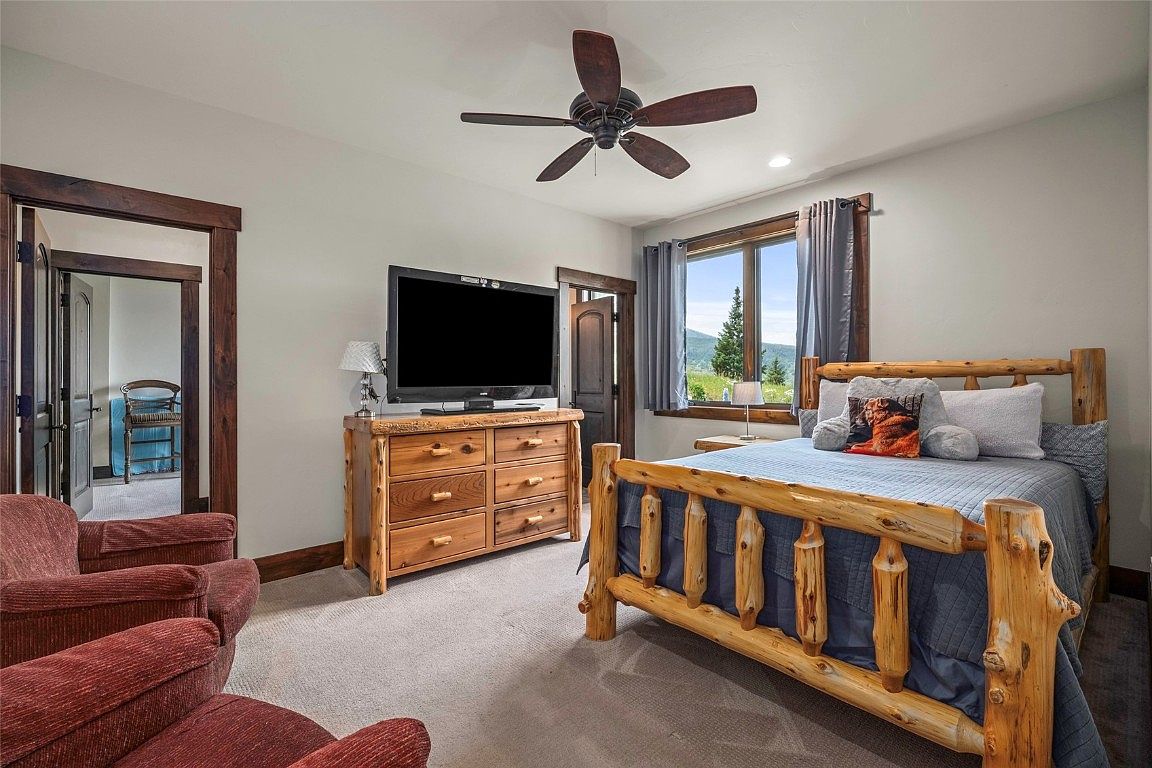This is a cozy bedroom featuring a rustic log bed with blue bedding and decorative pillows. A wooden dresser supports a large television, and a ceiling fan provides air circulation. The room has a warm and inviting atmosphere, enhanced by natural light from the window and comfortable seating.