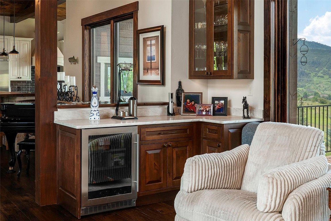 This interior shot showcases a cozy living room area featuring a built-in bar with wooden cabinetry and a stainless steel beverage cooler. A comfortable armchair sits nearby, inviting relaxation, while a window offers a glimpse of the outdoor landscape. The room exudes a warm and inviting atmosphere, perfect for entertaining or unwinding.