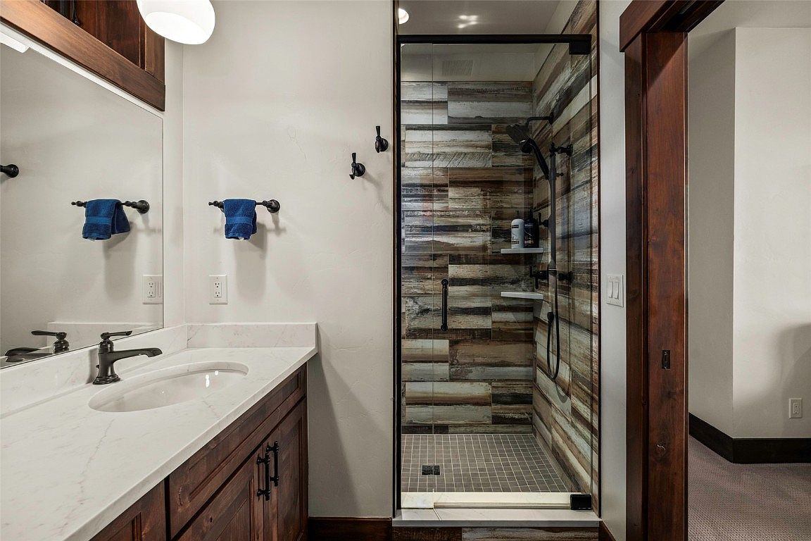This is a well-appointed bathroom featuring a wooden vanity with a white countertop and an oval sink. A framed mirror hangs above the vanity, and a glass-enclosed shower with wood-look tile and a gray tile floor is visible. The bathroom has a modern rustic style with dark hardware and warm wood tones.